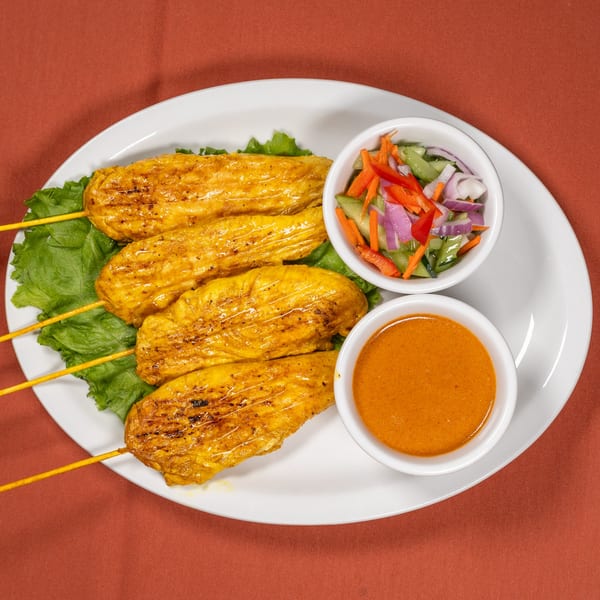 Four chicken satay skewers with peanut sauce and cucumber salad on a white plate.