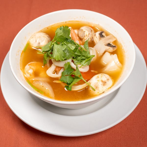A bowl of Tom Yum soup with shrimp, squid, mushrooms, and cilantro on a white plate.