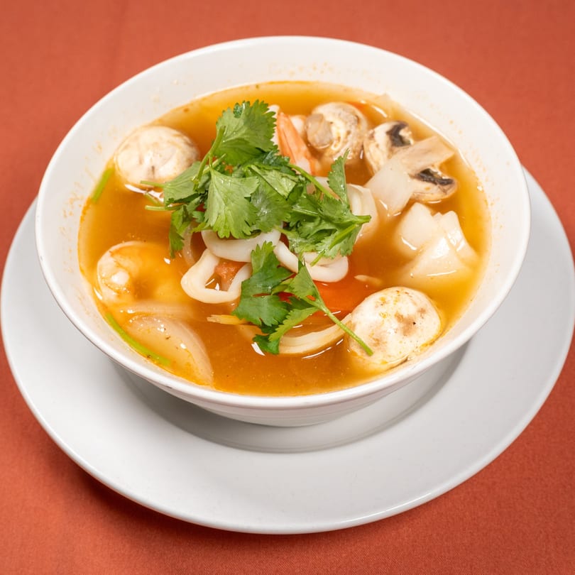 A bowl of Tom Yum soup with shrimp, squid, mushrooms, and cilantro on a white plate.
