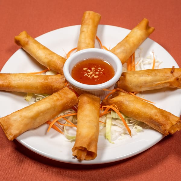 Golden-fried Thai spring rolls arranged around a small bowl of sweet chili dipping sauce on a white plate.