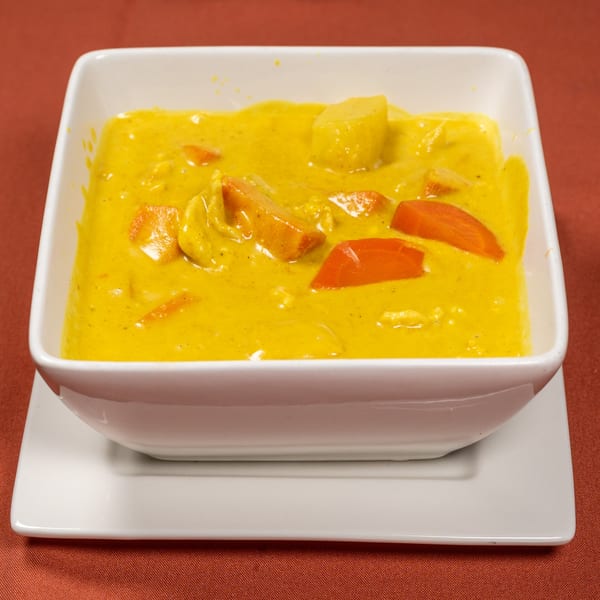 A white square bowl of yellow Thai curry with carrots and potatoes on a red tablecloth.